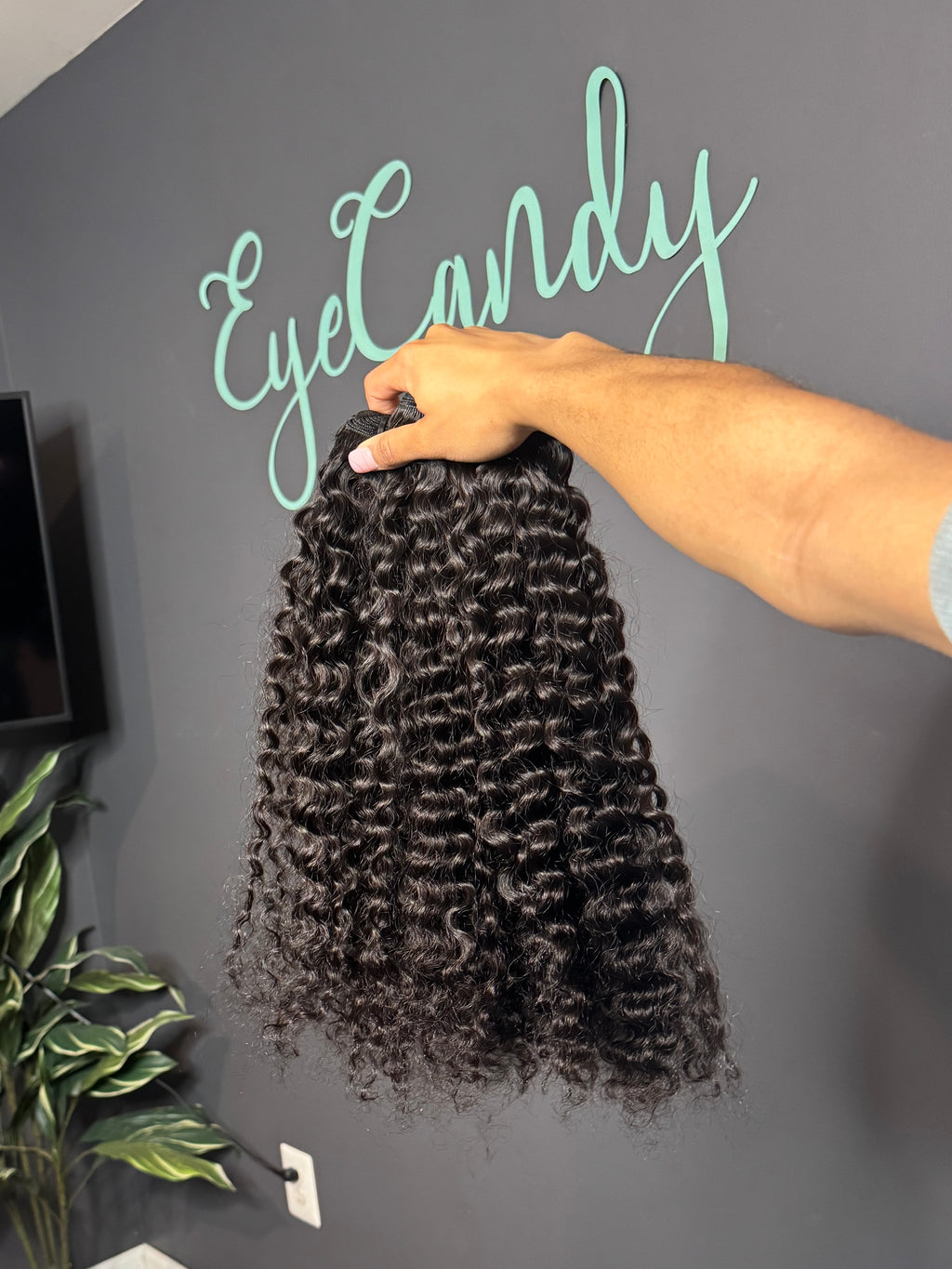 Person holding a bundle of Burmese curly hair extensions with 'EyeCandy' branding on a gray wall.