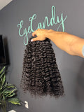 Person holding a bundle of Burmese curly hair extensions with 'EyeCandy' branding on a gray wall.