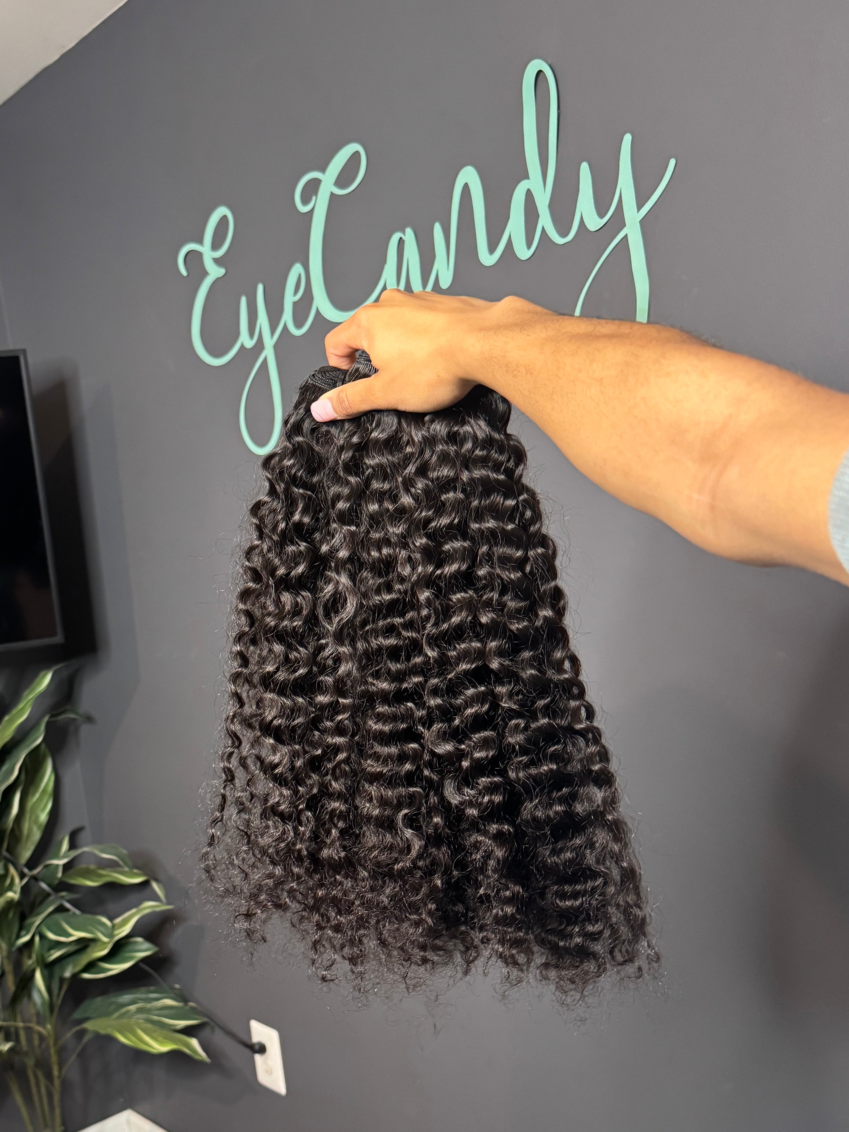 Person holding a bundle of Burmese curly hair extensions with 'EyeCandy' branding on a gray wall.